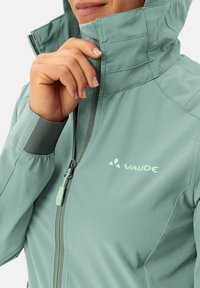Light green, soft-shell jacket with a high collar, front zipper, and elastic cuffs. Features a logo on the chest and textured fabric.
