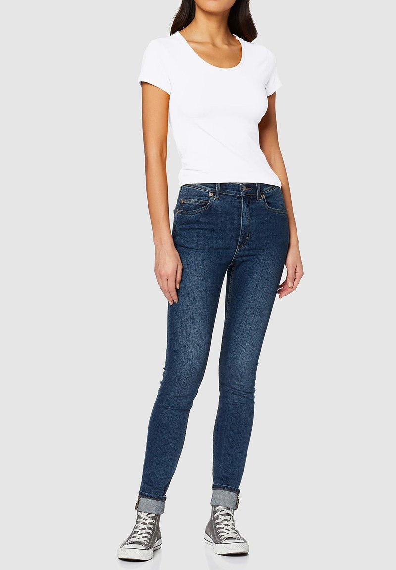 Jean skinny bleu foncé avec ceinture et poches avant, associé à un t-shirt ajusté blanc et des baskets montantes grises.