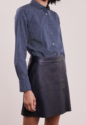 Mujer con camisa azul marino de lunares abotonada y metida dentro de una minifalda negra de cuero, de pie frente a un fondo liso.