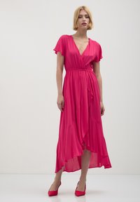 Femme avec des cheveux blonds courts portant une robe portefeuille rose vif avec des manches flottantes et des talons roses assortis, se tenant devant un fond blanc uni.