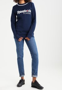 Marinblå Reebok-tröja med vit logotyp och röda accenter, kombinerad med ljusblå jeans och svarta sneakers. Slät tygkvalitet, avslappnad passform.