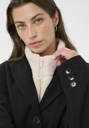 Schwarze Wollmischung Jacke mit einem krämfarbenen Kunstschafsfellkragen, Frontreißverschluss und drei schwarzen Knöpfen an den Bündchen. Geschmeidige Textur und taillierte Passform.
