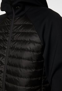 Svart vadderad jacka med vadderade frampaneler, släta ärmar av tyg och hög krage med dragkedja.