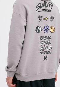 Grijze sweatshirt met gedurfde zwarte en kleurrijke grafische ontwerpen, inclusief tekst en symbolen. Zachte stof, losse pasvorm, geribbelde manchetten.