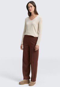 Beige Langarmshirt mit V-Ausschnitt, kombiniert mit braunen hochgeschneiderte, weit geschnittenen Hosen; Schuhe in einem helleren Tan mit Schnürdetails.