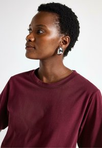 Sølvmetalliske øreringe med oval form, båret med en bordeaux farvet t-shirt med rund hals. Kort, tekstureret sort hår. Tæt profiludsigt.