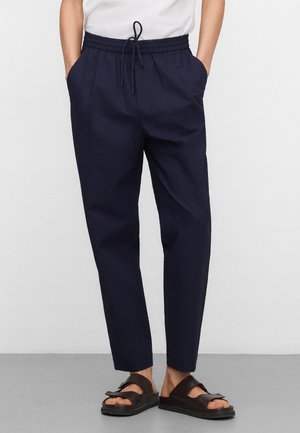 Persoon draagt marineblauwe, gestructureerde broek met trekkoord en zijzakken, gecombineerd met zwarte slides, op een grijze vloer tegen een witte muur.