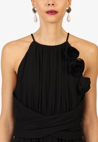 Schwarzes Kleid mit plissierter Textur, Halterneck-Ausschnitt und drei Stoffrosen auf der linken Schulter; verfügt über eine geraffte Taille und dünne Träger.