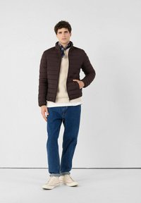 Donkerbruine pufferjas met hoge kraag, gestructureerd oppervlak en zijzakken, gedragen over een crèmekleurige trui en blauwe spijkerbroek, gecombineerd met lichte sneakers.