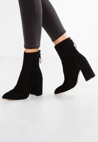 Bottines en daim noir avec un bout pointu, un talon bloc haut et un zip sur le côté. Texture lisse avec un design épuré.