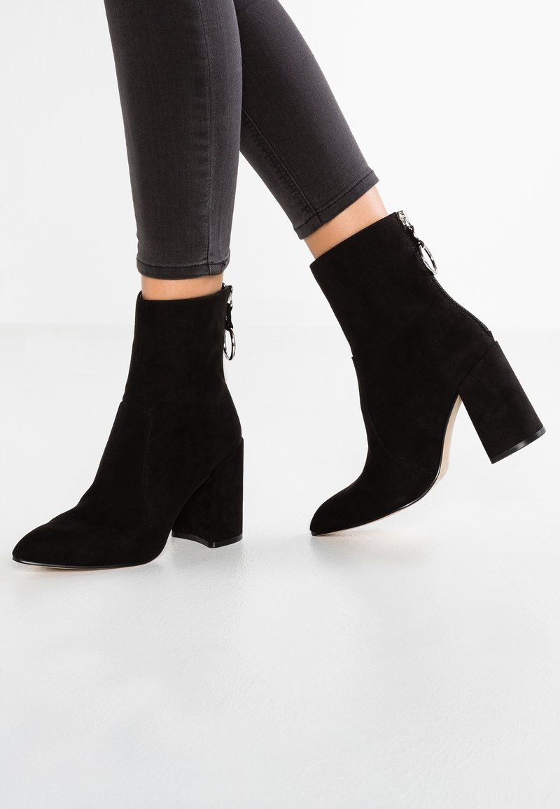 Bottines en daim noir avec un bout pointu, un talon bloc haut et un zip sur le côté. Texture lisse avec un design épuré.
