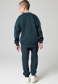 Tamsiai žalia palaidinė su tekstūrinėmis audinio savybėmis ir sutrauktomis rankovėmis, derinama su atitinkamomis kelnių kelnėmis. Baltos sportbačiai užbaigia aprangą.