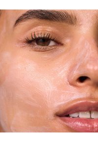 Nærbilde av et ansikt med en gjennomskinnelig, kremet ansiktsmaske påført. Jevn hudtekstur, mørke øyevipper og et svakt smil er synlig.