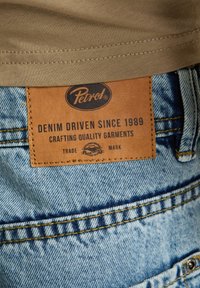 Pantaloncini di jeans con una tintura azzurra chiara, con una etichetta in pelle marrone chiaro impressa con "Petrol" e testo sulla storia e sulla qualità del marchio.