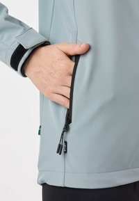 Main à peau claire avec des taches de rousseur dans la poche latérale zippée d'une veste bleu clair avec bordure noire et poignet ajustable.