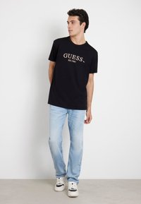 T-shirt en coton noir avec "GUESS" et "EST.1981" imprimé en marron clair. Assorti avec un jean bleu clair et des baskets blanches.