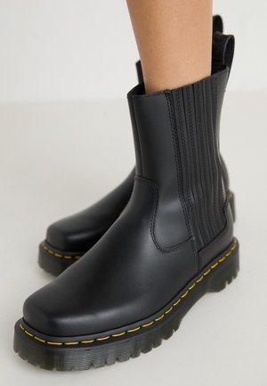 Bottes noires en cuir à la cheville avec coutures jaunes, panneaux latéraux élastiques et semelles épaisses en caoutchouc, portées par une personne debout sur une surface claire.