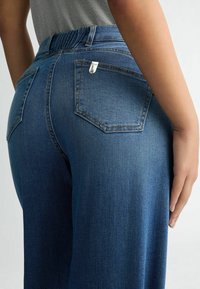 Jean en denim bleu foncé à taille haute, avec deux poches arrière et un petit badge de logo argenté sur la ceinture. Texture lisse.