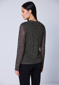Kvinde med langt mørkt hår iført en sort langærmet gennemsigtig top med små hvide blomsterprint og sorte bukser, stående sidevendt mod hvid baggrund.