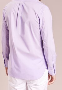 Man med en ljuslila långärmad skjorta och vita byxor sedd bakifrån mot en enkel beige bakgrund.