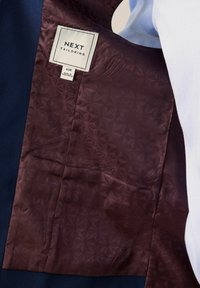 Saco de traje azul oscuro con un forro burdeos estampado, con una etiqueta que dice "NEXT Tailoring" y la información de la talla.