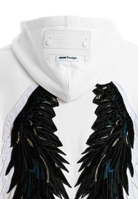 Sudadera blanca con intrincados diseños de alas bordados en negro y azul en el frente, y una etiqueta minimalista en la parte posterior.
