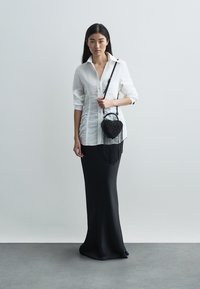 Λευκό, πουκάμισο με κουμπιά και διακριτή υφή, συνδυασμένο με εφαρμοστή μαύρη μακριά φούστα· μαύρη τσάντα σε σχήμα καρδιάς με λεπτομέρειες κρόσσια.