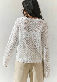 Pull en tricot blanc avec un motif en chevron, manches longues et ourlet festonné. Porté sur un pantalon beige avec des coutures décoratives.