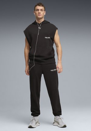 Schwarzer ärmelloser Hoodie mit Fronttasche und "PUMA CREW"-Aufdruck. Kombiniert mit passenden schwarzen Hosen und neutralfarbenen Sportschuhen.