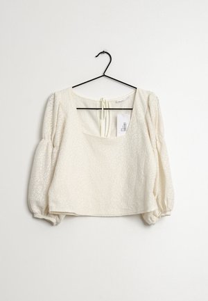 Blouse blanche texturée avec un col carré, des manches bouffantes et une fermeture à nouer dans le dos. Suspendue à un crochet noir.