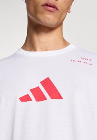 Valge puuvillane t-särk, millel on esiküljel silmatorkav punane graafika, millel on kolm kaldus triipu, koos punase trükitud tekstiga.