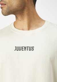 Svetlo béžové bavlnené tričko s okrúhlym výstrihom a krátkymi rukávmi, so slovom "JUVENTUS" v tučných čiernych písmenách cez hruď.