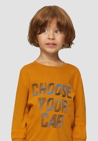 Oranje shirt met lange mouwen, voorzien van de opvallende tekst "KIES JE AUTO" in zwart en blauw. Zacht, katoenen materiaal met een losse pasvorm.