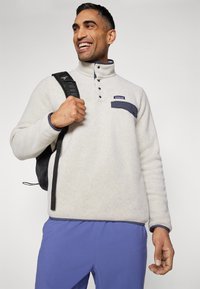 Pulover din fleece de culoare gri deschis, cu accente navy, guler înalt, trei butoane și un buzunar pe piept. Asortat cu pantaloni casual albaștri.
