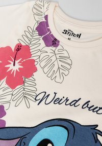 Valkoinen kangas, jossa on painettuja trooppisia lehtiä, purppuran ja punaisen hibiskuksen kukkia, osittainen sininen sarjakuvahahmo ja "Weird out" -teksti kaula-aukossa.