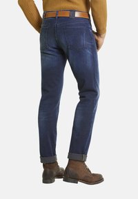 Jean en denim bleu foncé avec une coupe droite, présentant des poches arrière et des ourlets retroussés. Associé à des bottes brunes à lacets et une ceinture beige.