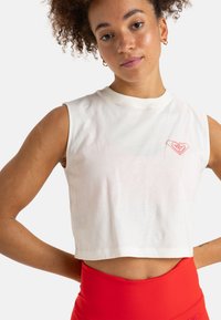 Cropped white sleeveless top made of cotton, featuring a small red heart design on the upper left. Paired with high-waisted red leggings.