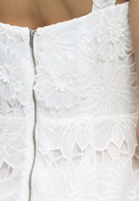 Λευκό φόρεμα με μεγάλα λουλουδάτα δαντελωτά σχέδια και κεντρικό ασημί φερμουάρ, φορεμένο από άτομο με γυμνούς ώμους.