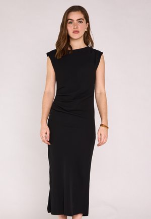 Femme aux longs cheveux bruns portant une robe midi noire sans manches, des boucles d'oreilles dorées et des bracelets marron, debout devant un fond clair uni.