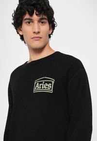 Fekete hőszigetelő pulóver, bordázott textúrával, széles nyakkivágással és bézs színű "Aries" logóval, amely a mellkason van hímezve.