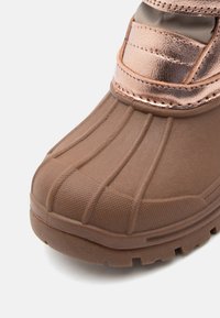 Barna vízálló csizma, bordázott gumitalppal, textúrált felsőrésszel és fémből készült bronz kiegészítővel a tetején. Puffertömörítéssel ellátott szegéllyel rendelkezik.