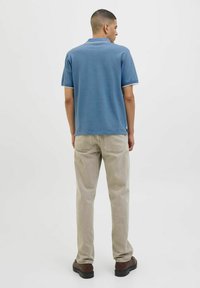 Kék póló klasszikus gallérral, rövid ujjú, fehér díszítéssel. Bézs kordbársony nadrág. Fekete cipők. Hátulnézet egy sima háttér előtt.