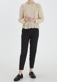 Blusa beige con motivo nero, collo e maniche volant, orlo peplum. Indossata con pantaloni neri a sigaretta e mocassini neri.