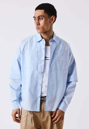 Jeune homme portant une chemise bleu clair boutonnée sur un t-shirt blanc à motif et un pantalon beige, regardant vers sa gauche.