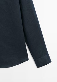 Dlouhý rukáv košile z tmavě modrého lněného materiálu s manžetou a viditelným stehem na bílém pozadí.