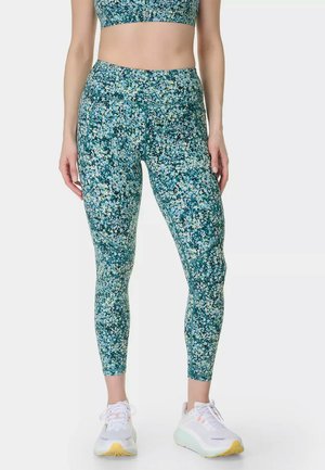 Femme portant un legging vert à motif floral, un crop top assorti et des baskets blanches, debout devant un fond uni.