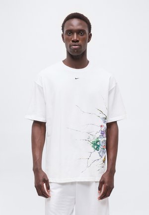 Mann trägt weißes Nike T-Shirt mit kleinem schwarzem Logo auf der Brust und buntem, rissigem Design unten rechts, steht vor schlichten weißem Hintergrund.