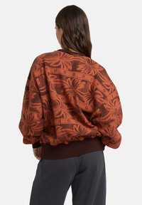 Przesadzony sweter w kolorze wypalonej pomarańczy z ciemnymi kwiatowymi wzorami. Posiada ściągacz na dole i przy mankietach, miękką fakturę oraz przytulny fason.