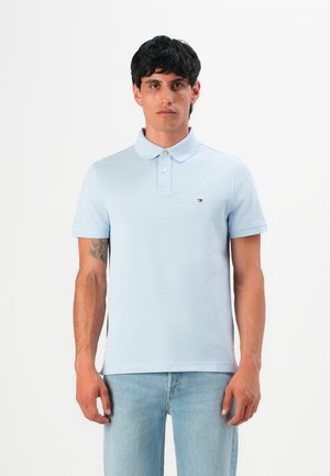 Mann in einem hellblauen Poloshirt und hellen Jeans, der vor einem schlichten weißen Hintergrund steht.