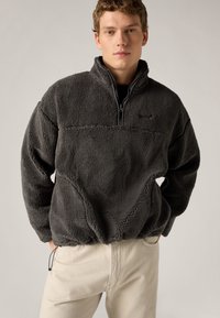 Pull-over en polaire grise avec une demi-fermeture éclair, surface texturée et coupe décontractée. Comprend un petit logo sur la poitrine et des ourlets élastiques.